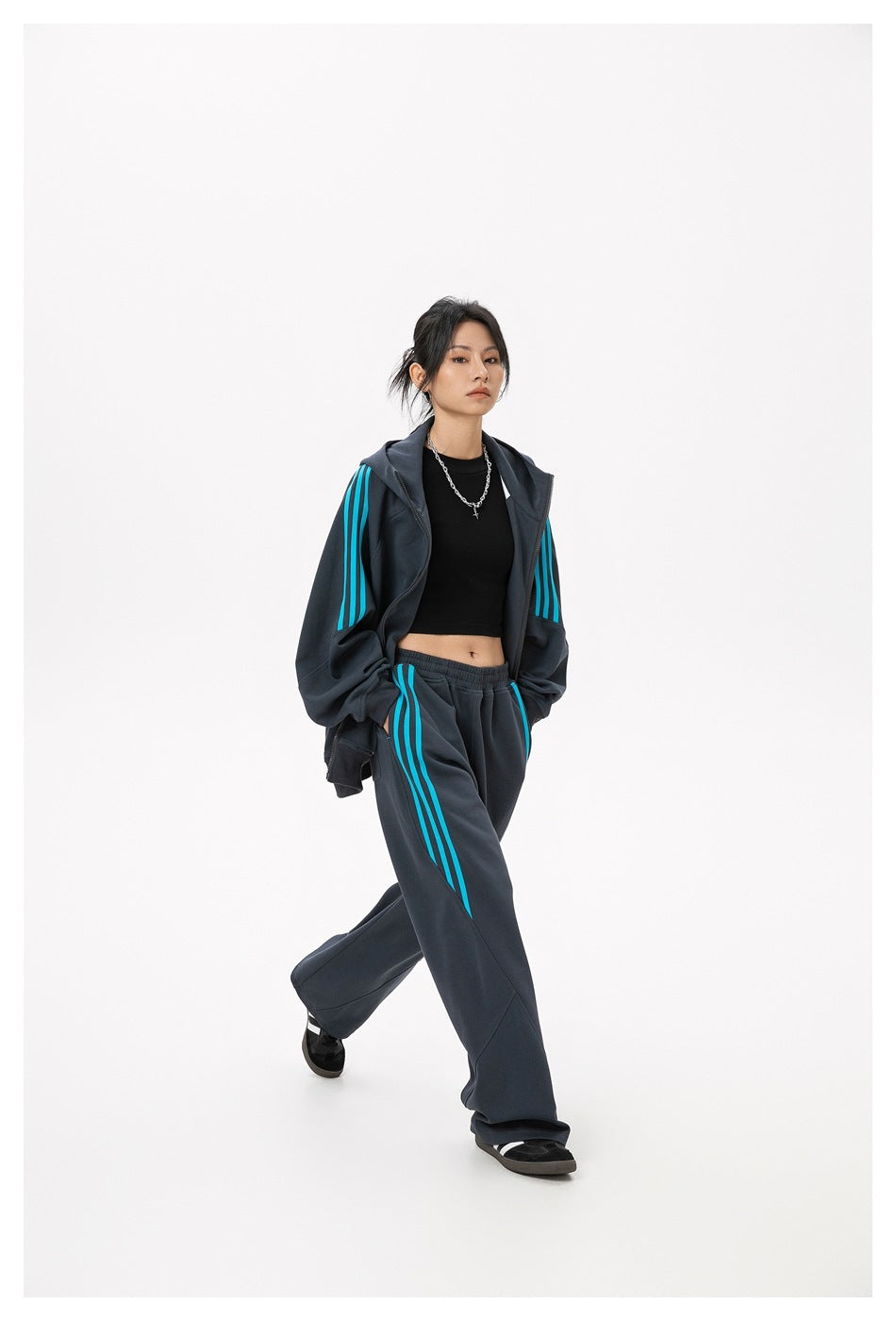 Model wearing an open charcoal hoodie and loose matching pants with bright cyan stripes and a black crop top.
