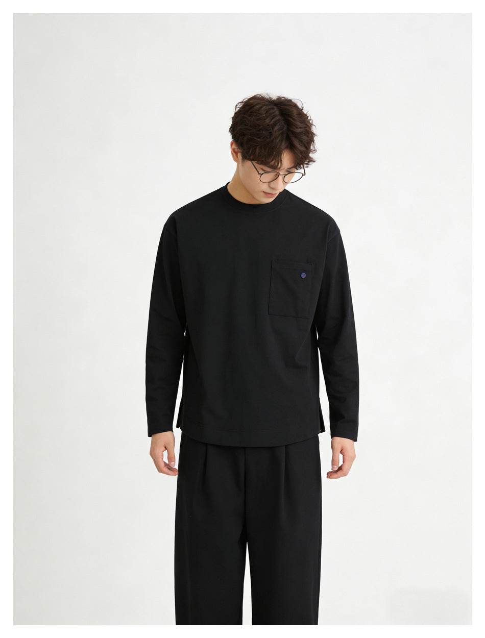 Model looking down while wearing a black long-sleeve crewneck shirt with a chest pocket and black trousers.