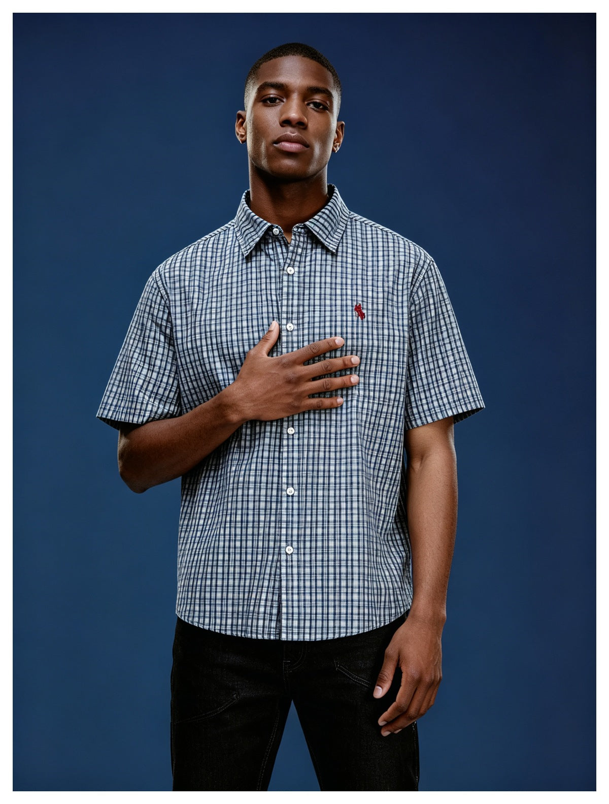 Man wearing a blue check short-sleeve button-up shirt with red chest logo and black pants against a navy background