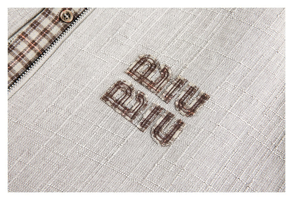 Embroidered lettering on cream textured fabric beside brown plaid trim and a button.