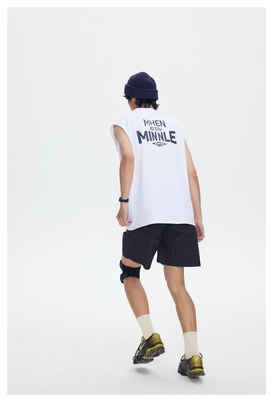 Model seen from behind wearing a white sleeveless top with navy back print, black shorts, beanie and sneakers