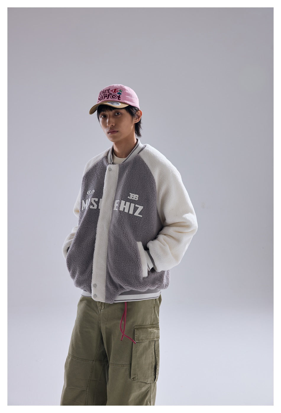 Standing model wearing a gray-and-cream snap-front jacket with chest lettering and khaki cargo pants.