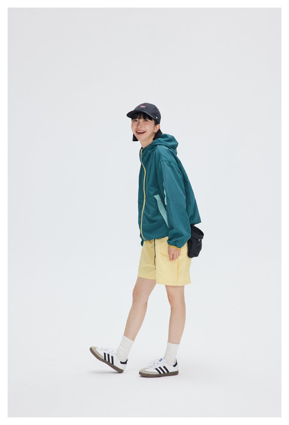 Side view of a model in a teal hooded zip jacket with yellow zipper and mint panel, with yellow shorts and a cap