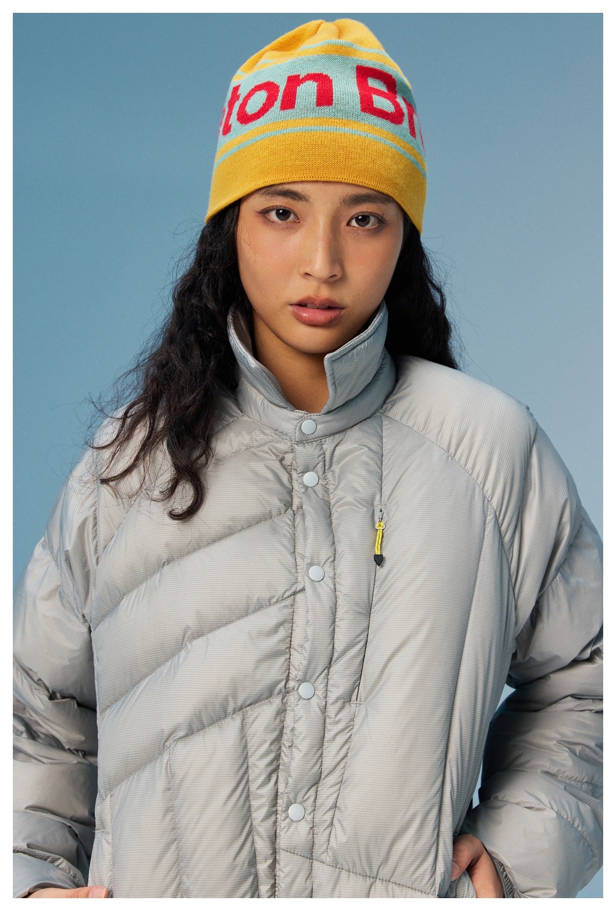 Model wearing a light gray quilted jacket with snap closures and a yellow knit beanie with red lettering.