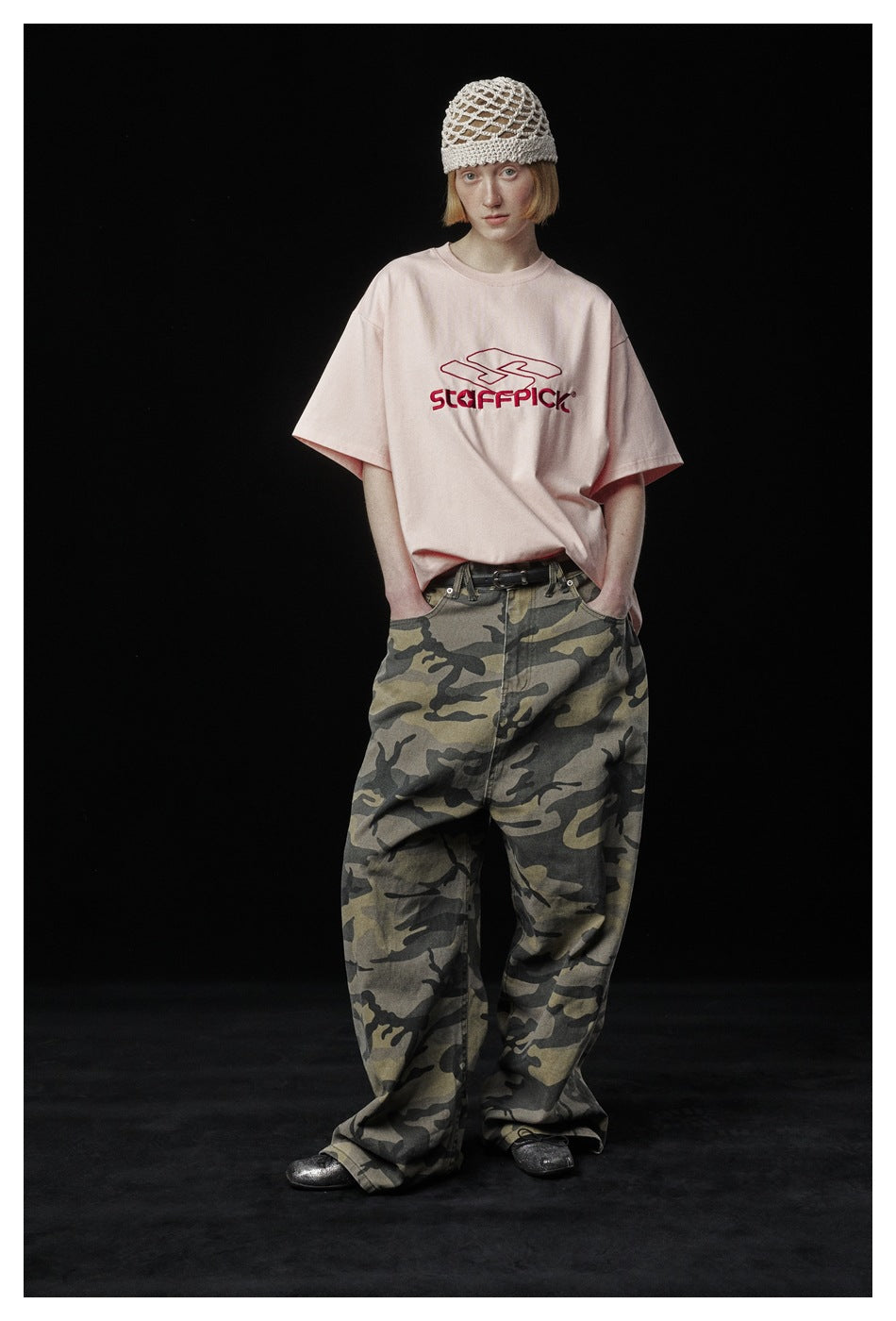 Model wearing a pale pink graphic T-shirt and loose camouflage trousers, standing with hands in pockets.