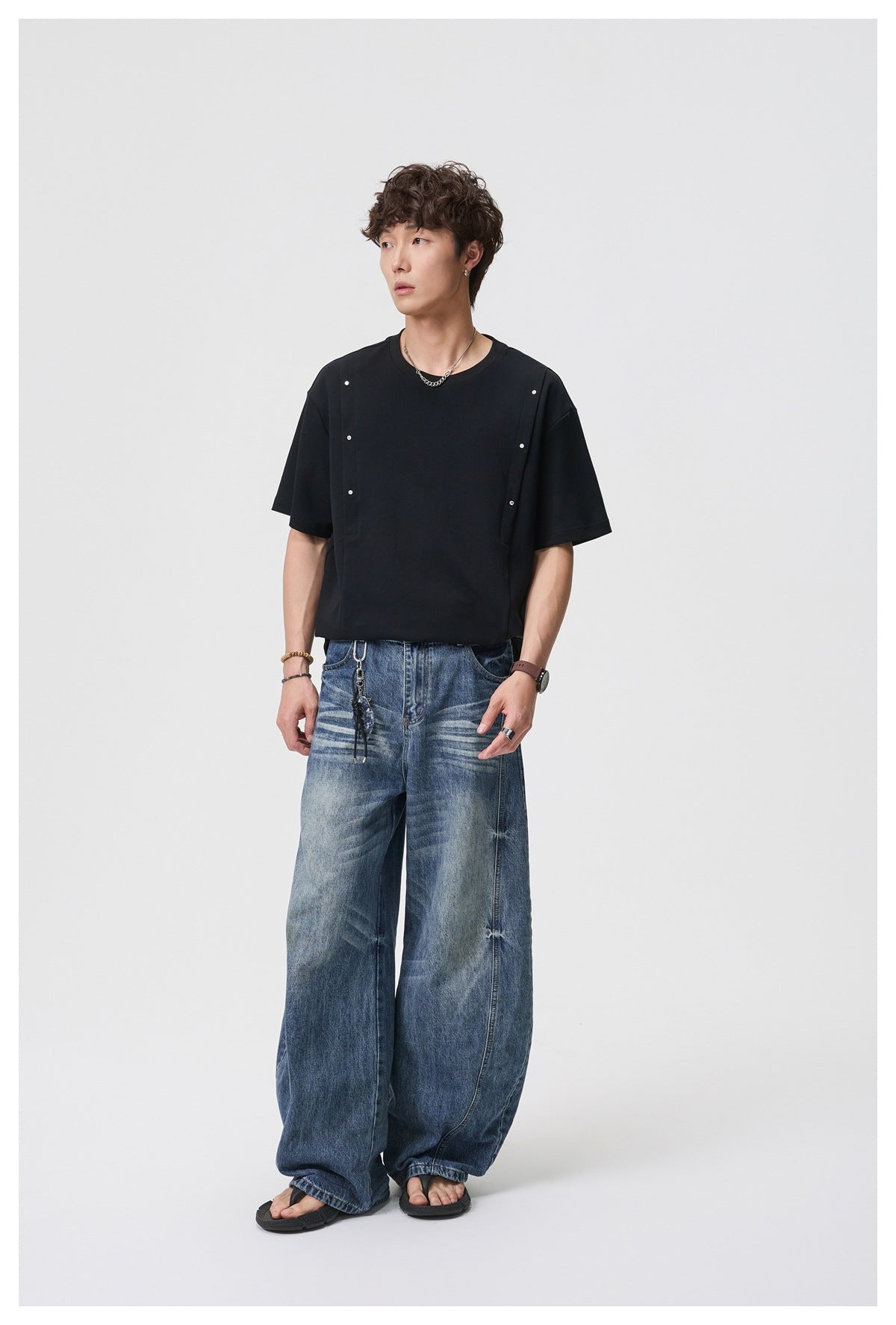 Man in a black rivet-detail T-shirt with wide-leg washed blue jeans and sandals, full-length view