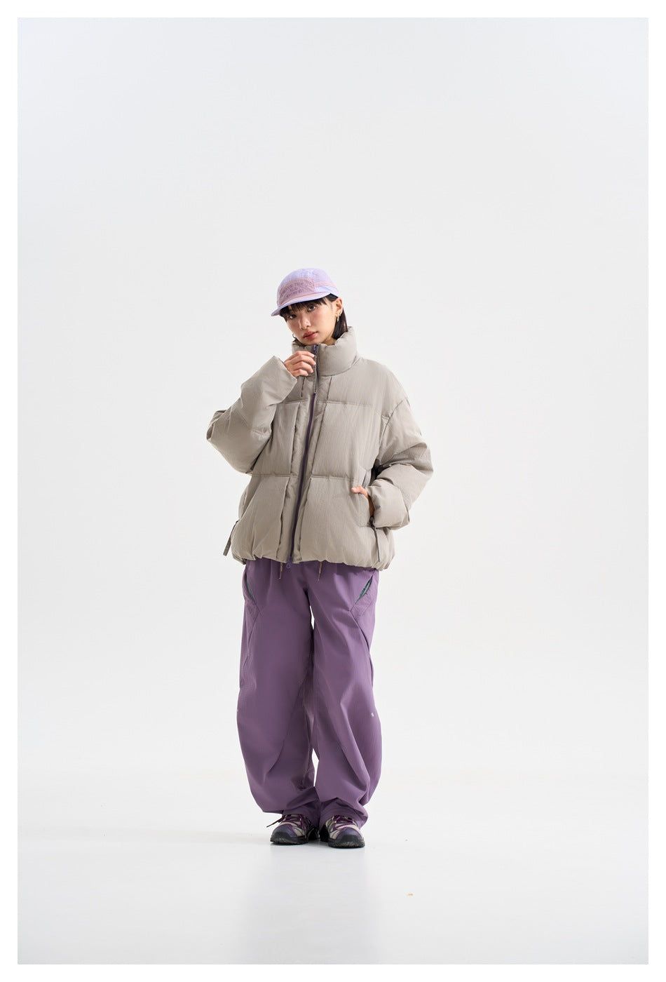 Full-length model in a light gray oversized puffer jacket, loose purple trousers, and a lavender cap.