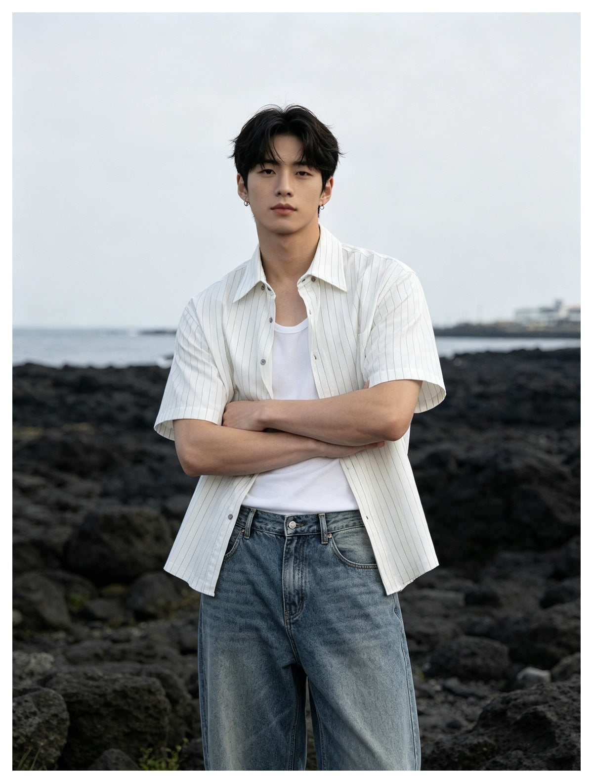 Man wearing white pinstripe short-sleeve shirt open over a white tank top and jeans, standing by dark rocks and sea