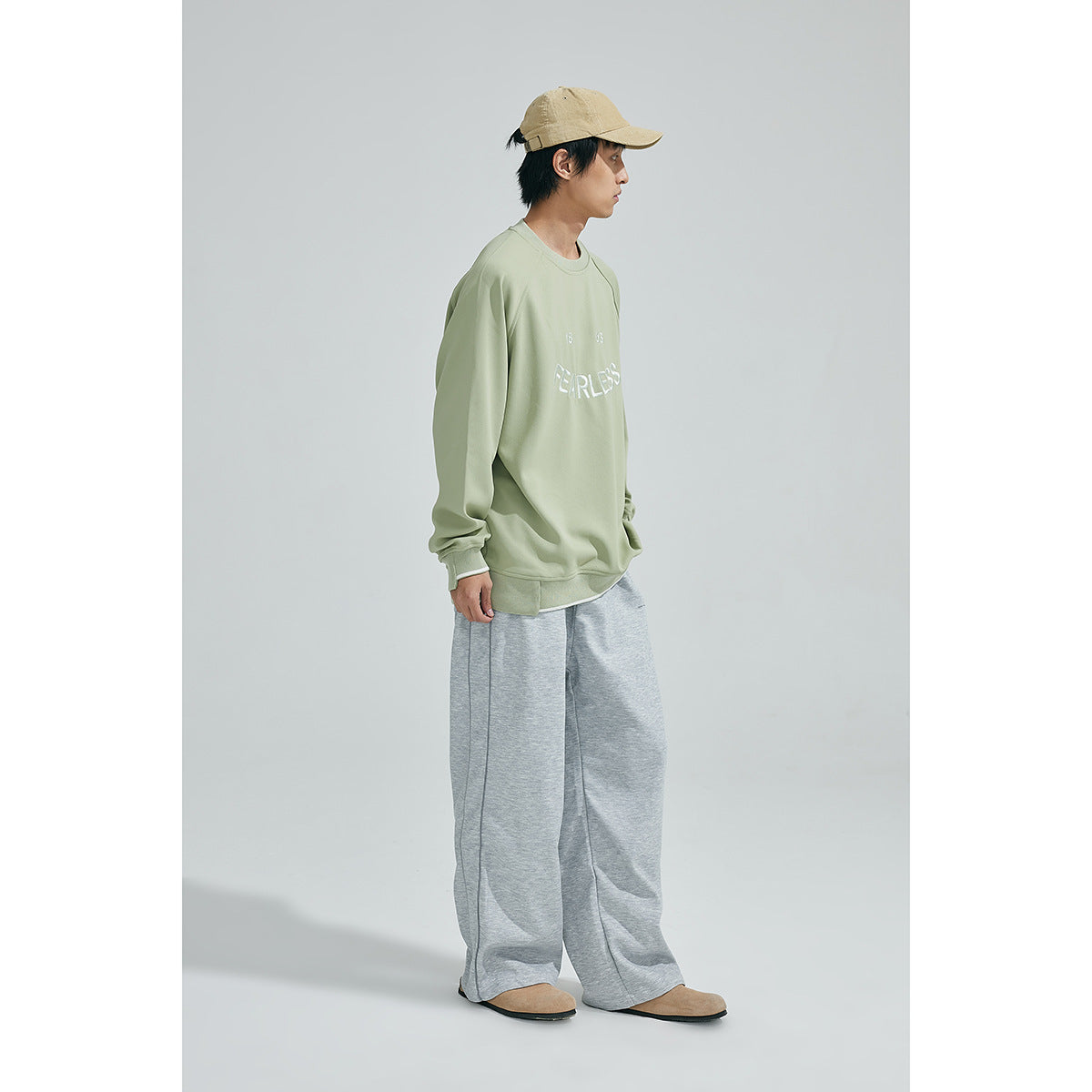 Model in profile wearing a sage green FEARLESS sweatshirt, loose gray pants, and a tan cap.