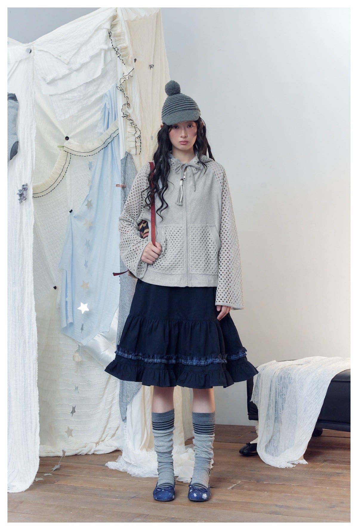 Model wearing gray mesh-sleeve zip hooded jacket with a navy tiered skirt, beanie and striped socks, front view