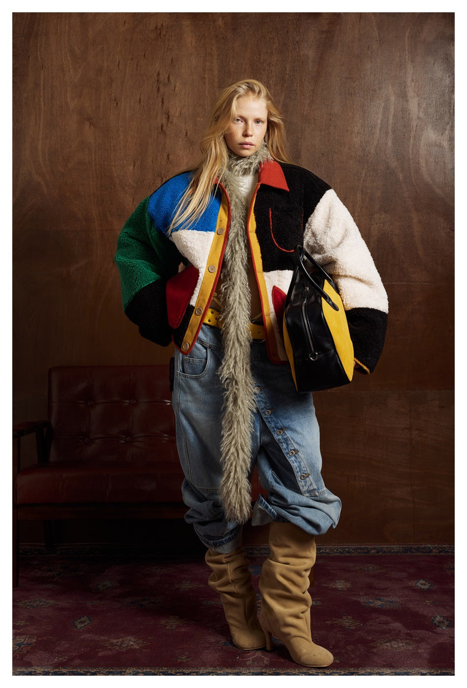 Model wearing the color-block fleece-like jacket open over a scarf, wide jeans and tan boots, holding a bag