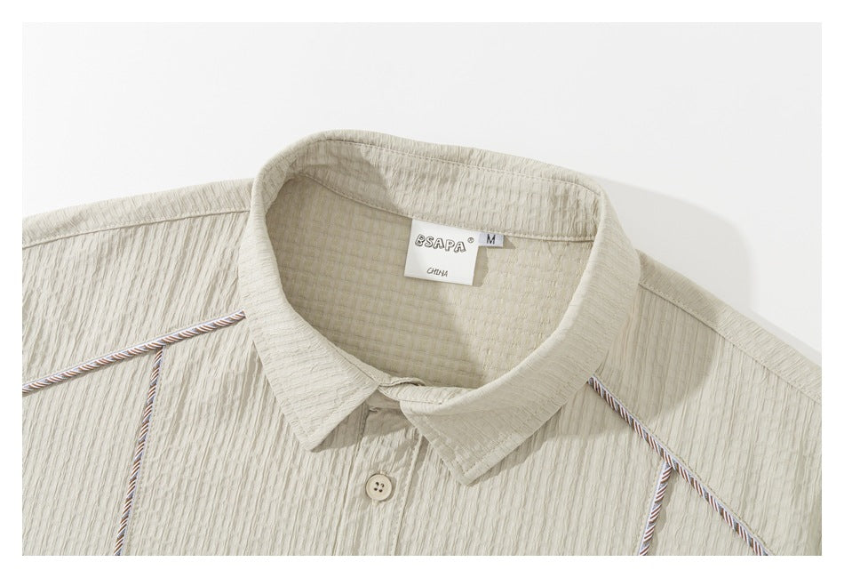 Close-up of a light beige shirt collar, textured fabric, button, contrast piping, and sewn-in neck label.