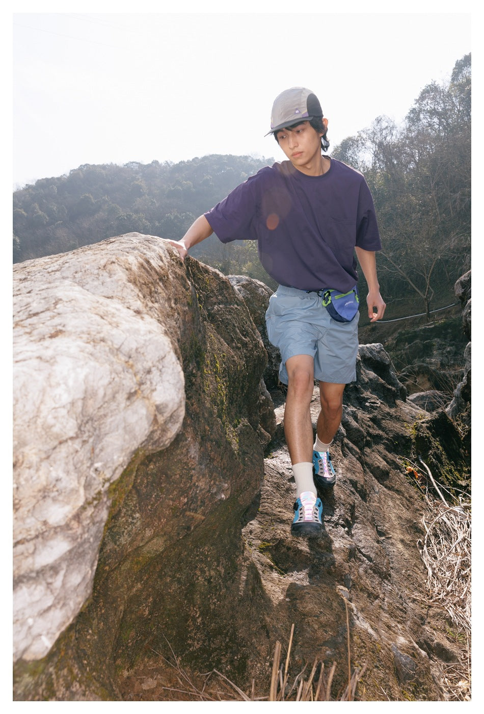 Model hiking in a purple oversized T-shirt and light blue shorts with a blue waist pack.