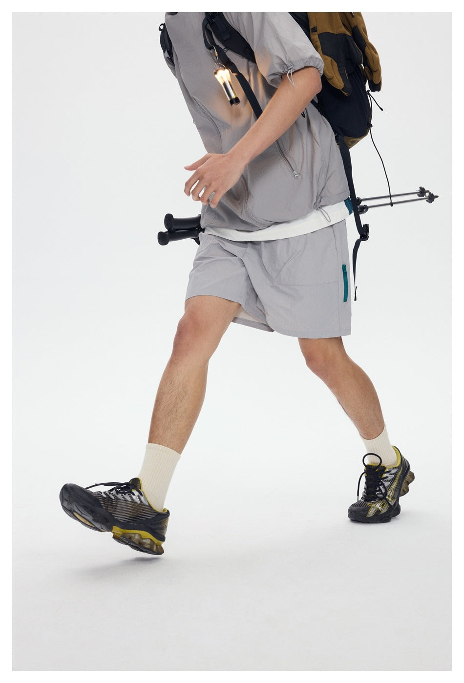 Cropped model walking in a light gray outdoor jacket and matching shorts, carrying a backpack and trekking poles.