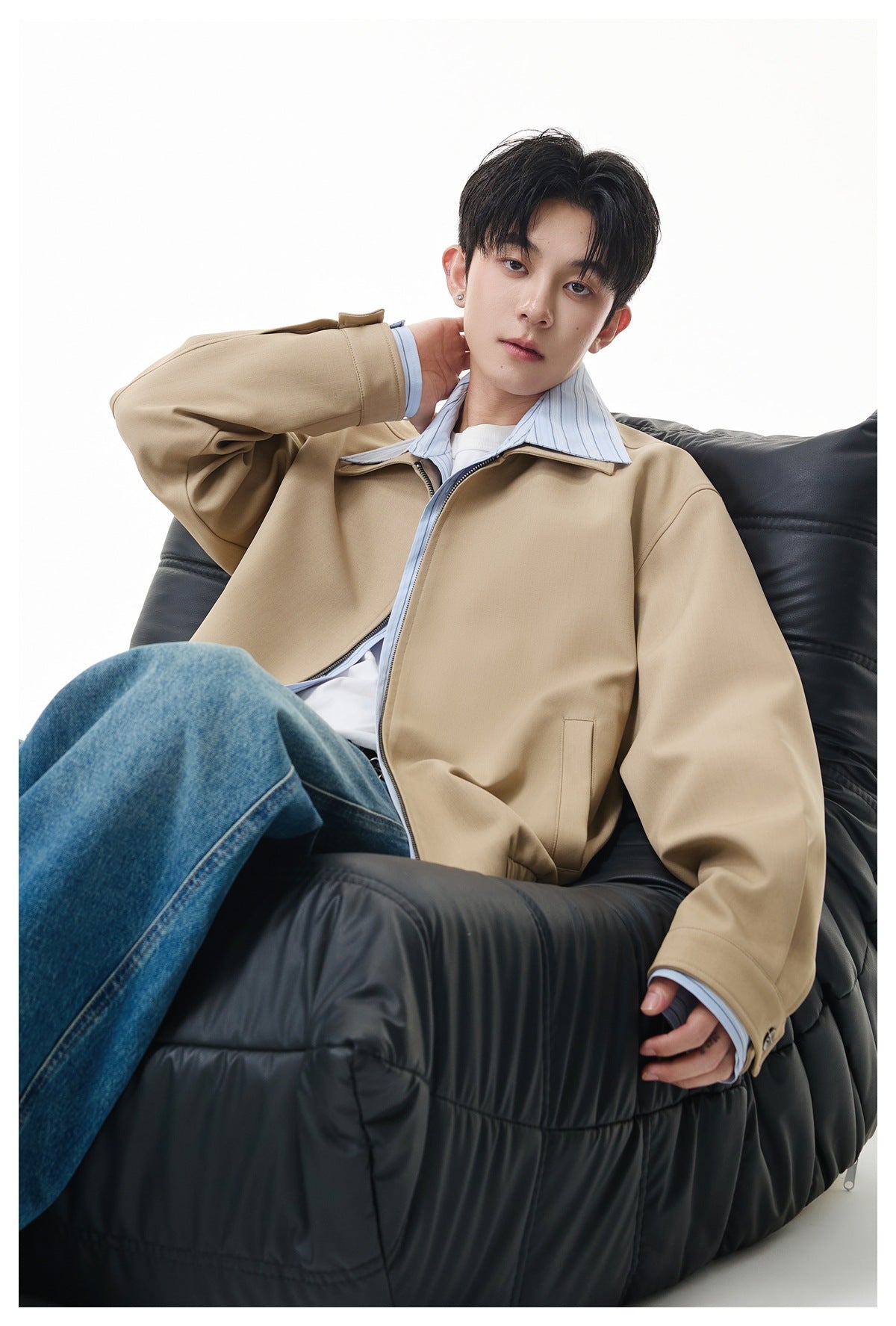 Model seated on a black puffy chair wearing a khaki zip jacket over a striped shirt, white tee and blue jeans