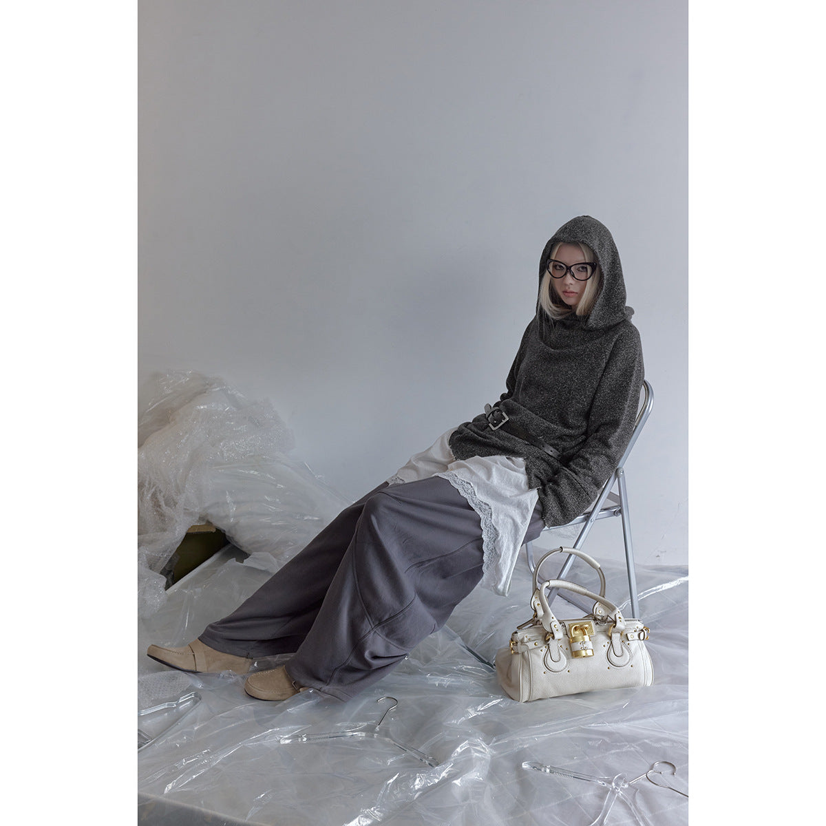 A seated model wears a charcoal hooded top with a black belt, layered white lace-trim shirt and loose gray pants.