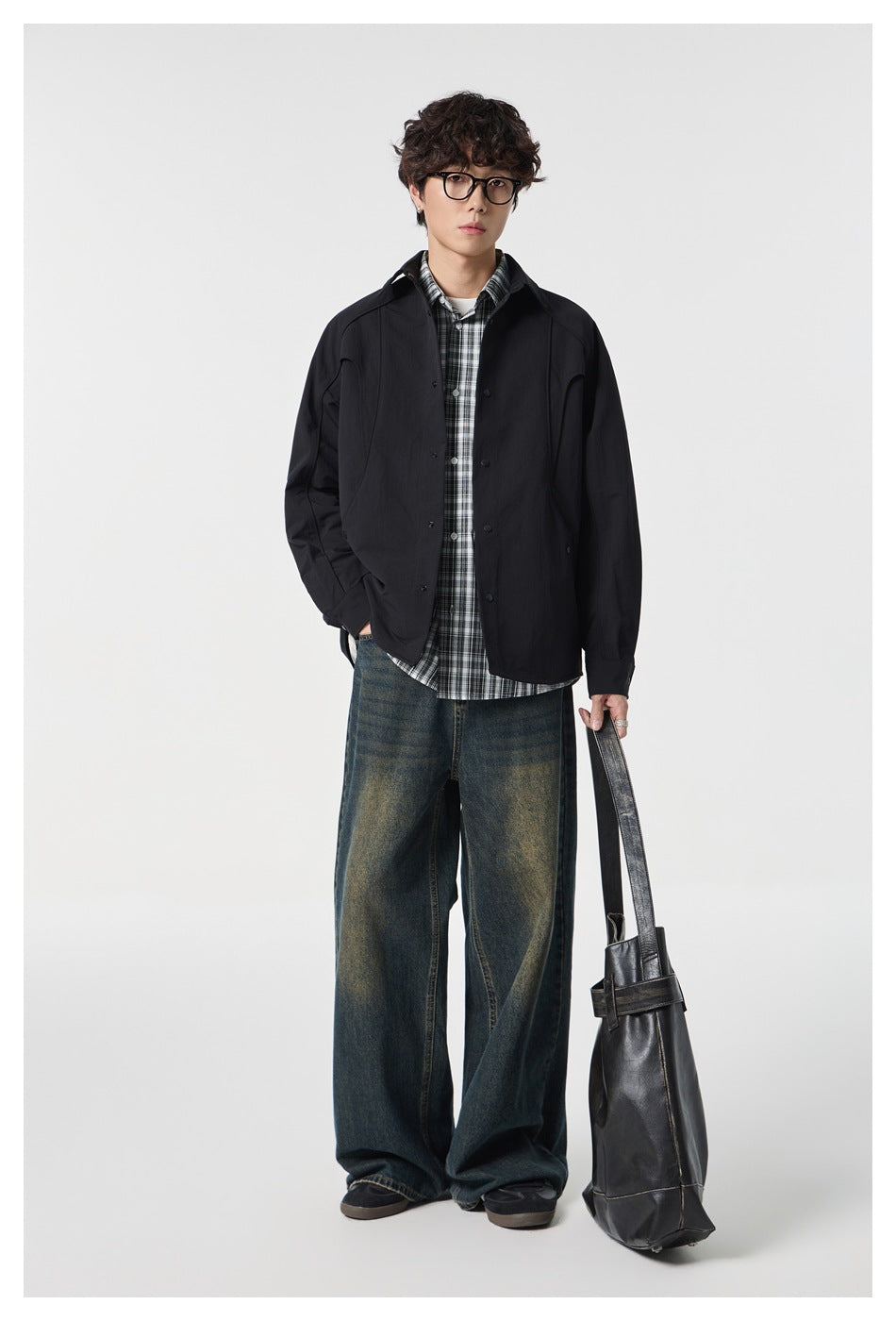 Full-length view of model in black shirt jacket over plaid shirt with wide-leg jeans, holding a bag