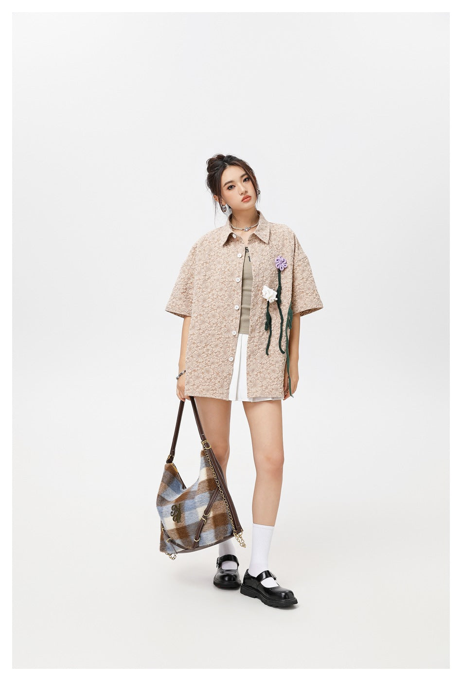 Model wearing a beige floral-textured button-up shirt with flower appliqués over a white skirt, shown with a shoulder bag.