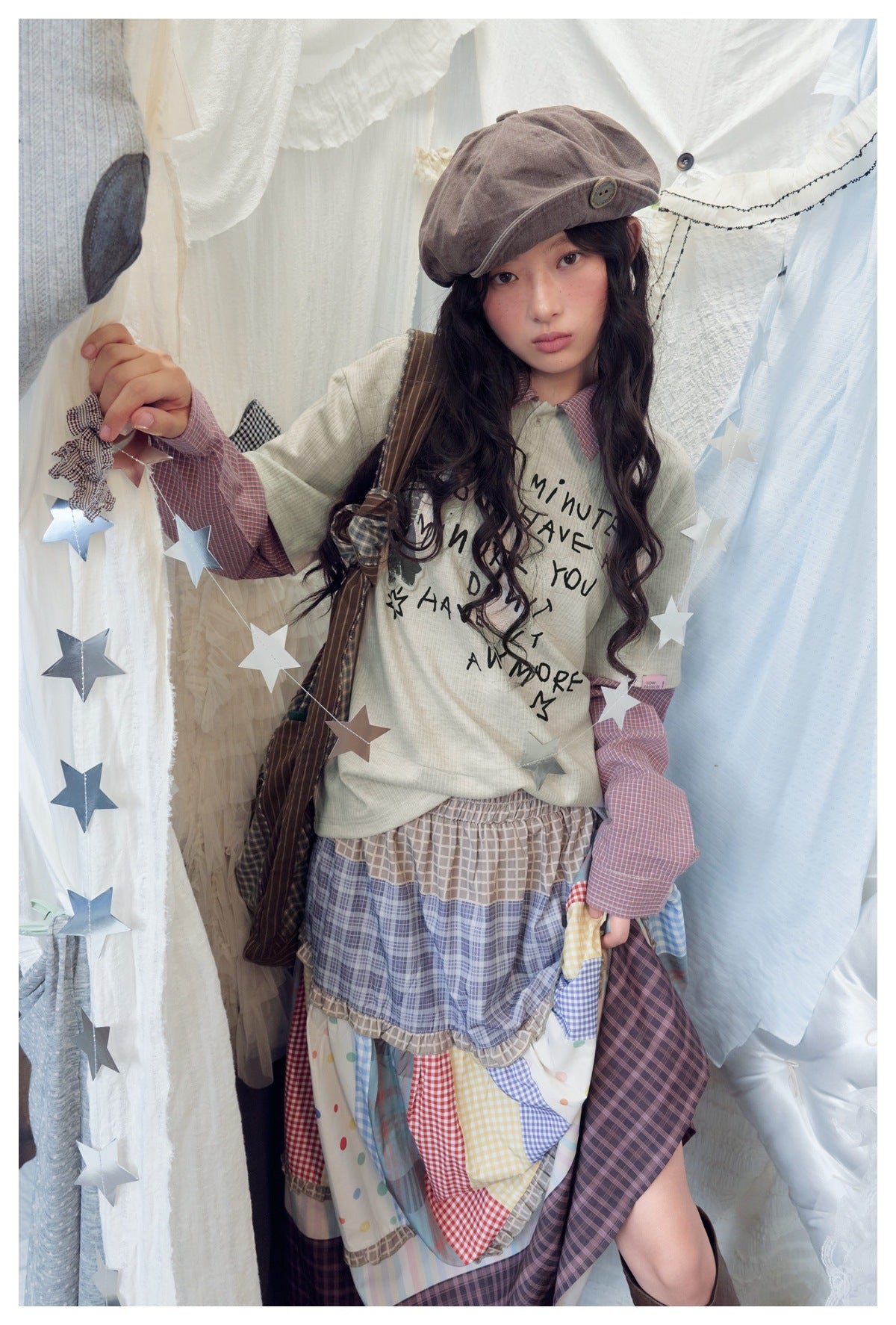 Model wearing apricot polo-collar top with pink check sleeves, a beret and a patchwork check skirt, holding a star garland