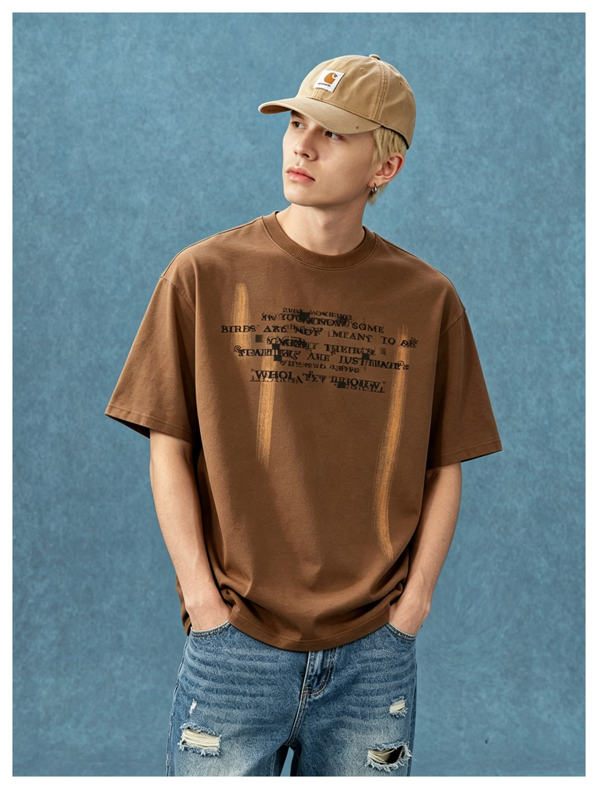 Young man wearing a brown oversized T-shirt with distorted text print and jeans against a blue backdrop