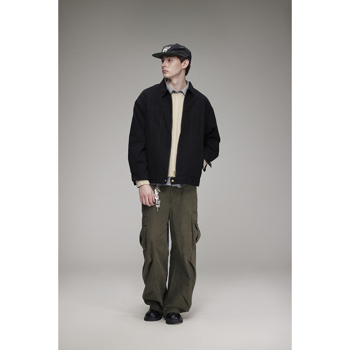 Full-length view of a model wearing a black jacket, cream sweater, checked shirt, black cap, and loose olive cargo pants.