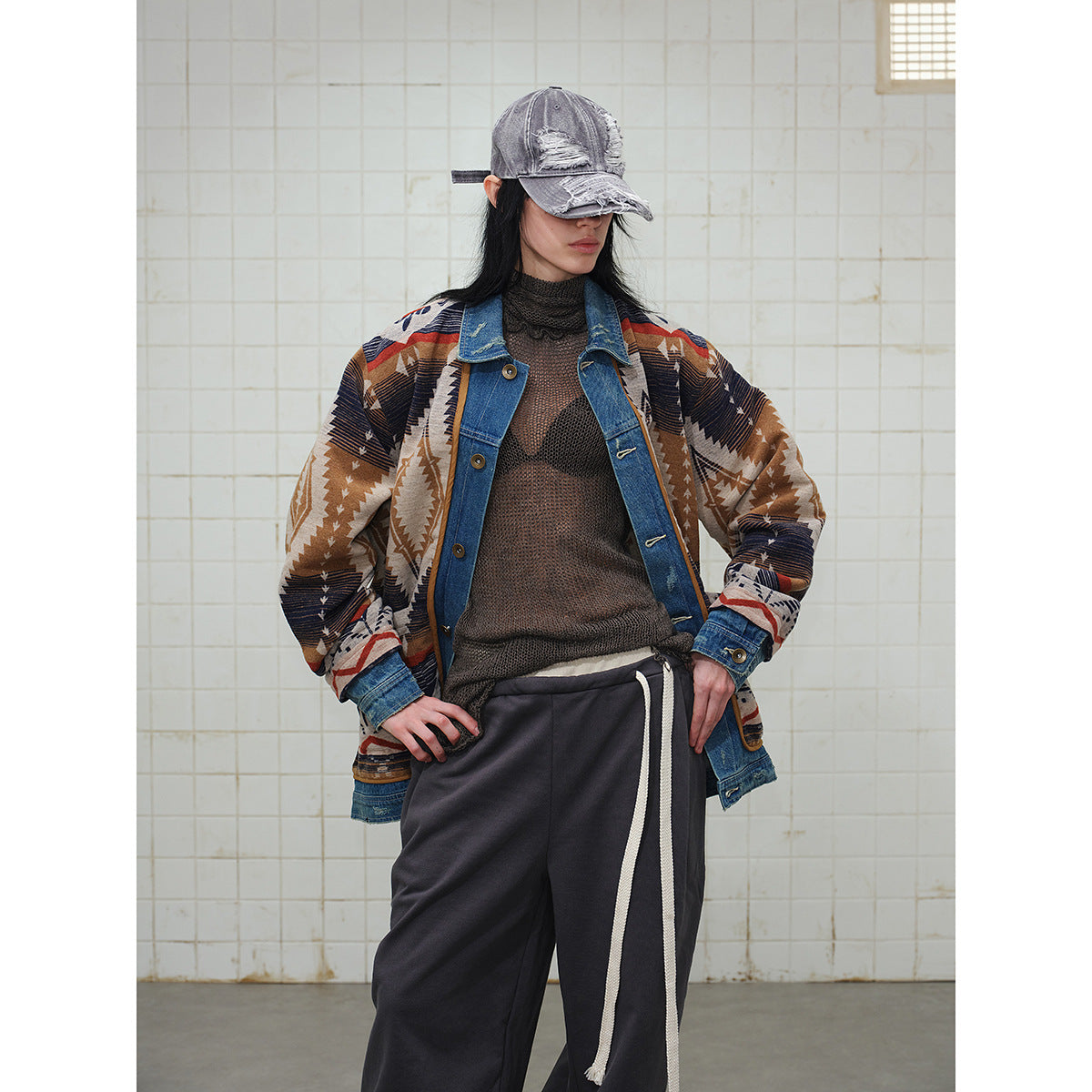 Model wearing an open geometric-pattern jacket with denim trim, a dark sweater, charcoal drawstring pants and a distressed