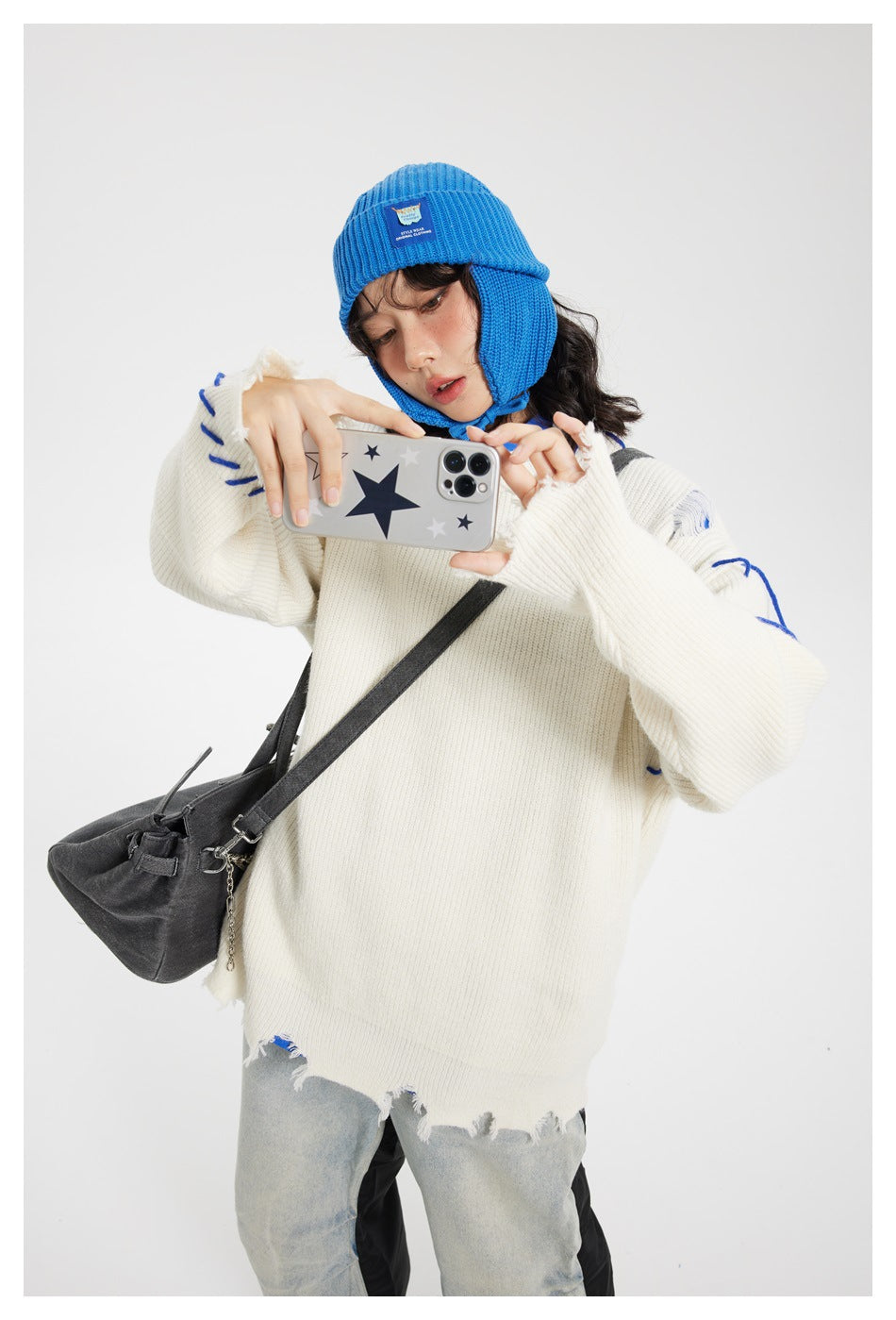 Model in an ivory distressed sweater with blue accents and a bright blue earflap beanie, holding a phone for a selfie.