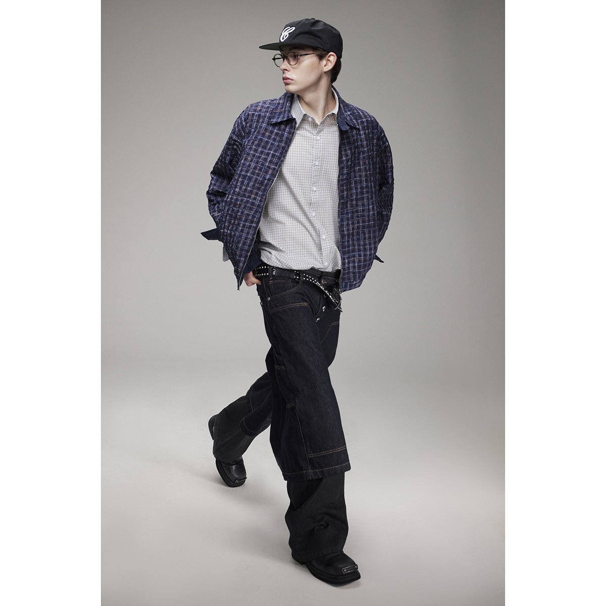 Model in a navy plaid jacket, checked shirt, black cap, and loose dark jeans, shown walking.