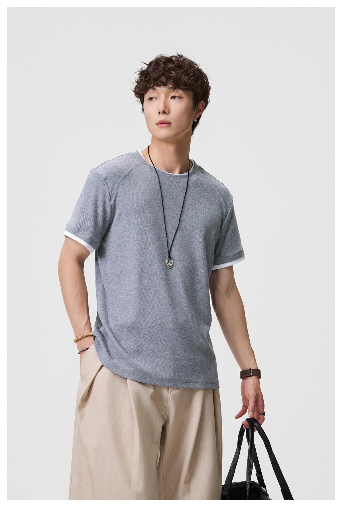 Man wearing gray layered-look short-sleeve T-shirt with beige pleated trousers, holding a black bag, angled pose