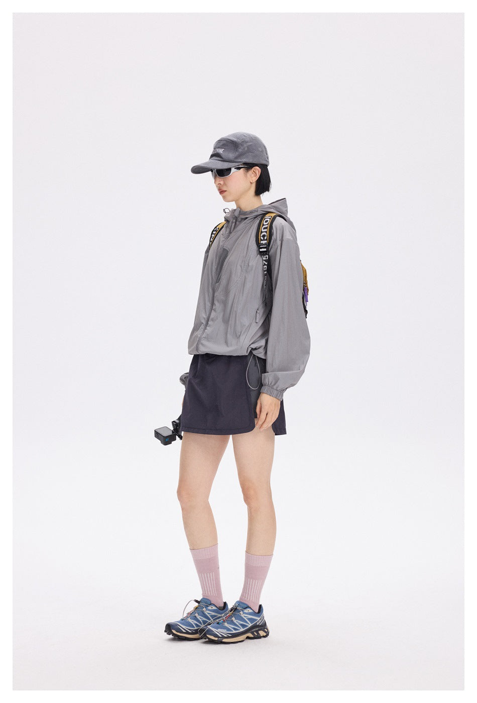Side view of a model wearing a gray hooded jacket, dark navy shorts, a cap and a backpack.