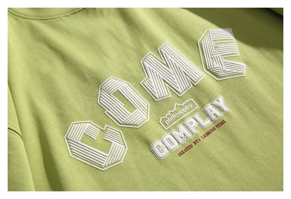 Close-up of the white embroidered English lettering and graphic on a light olive-green T-shirt.