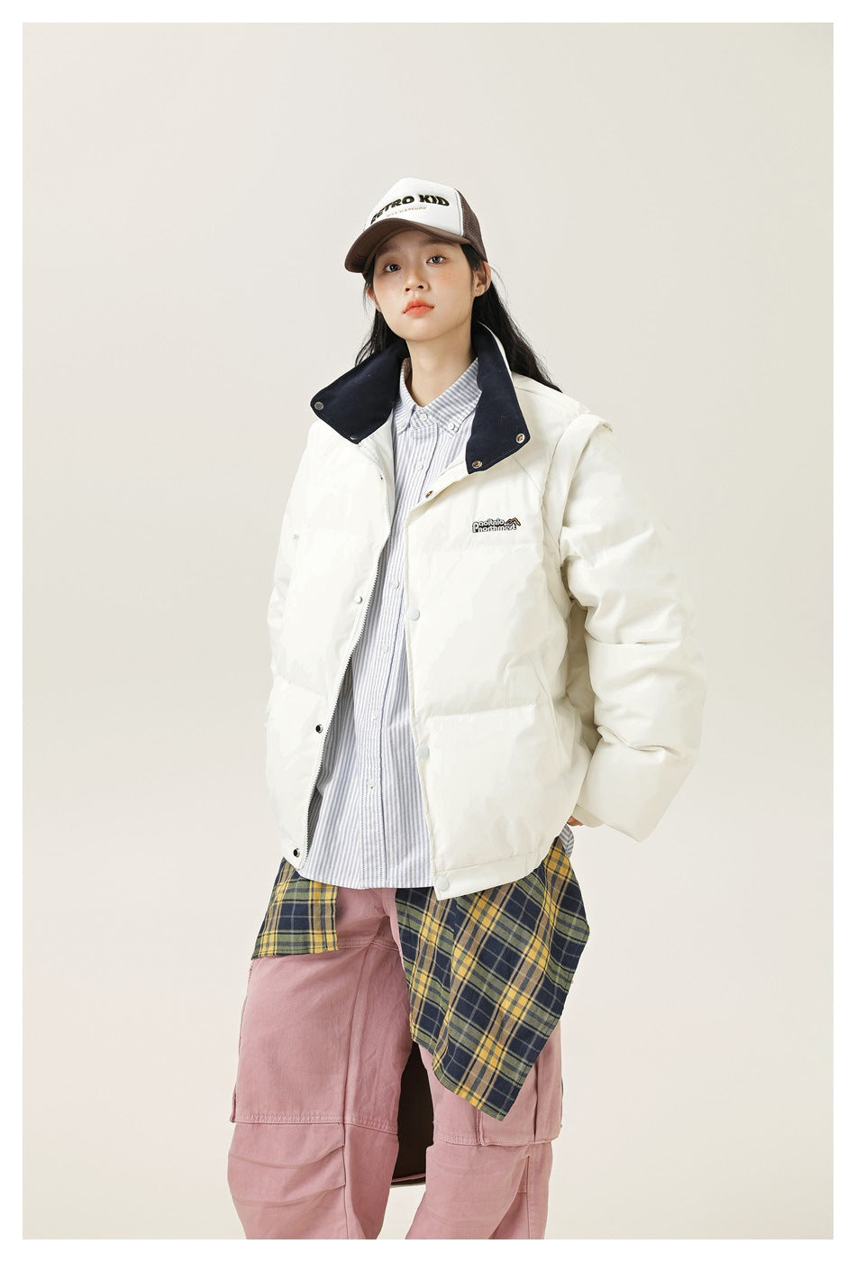 A model wears a white padded jacket with a navy collar over a striped shirt and a white-and-brown cap.