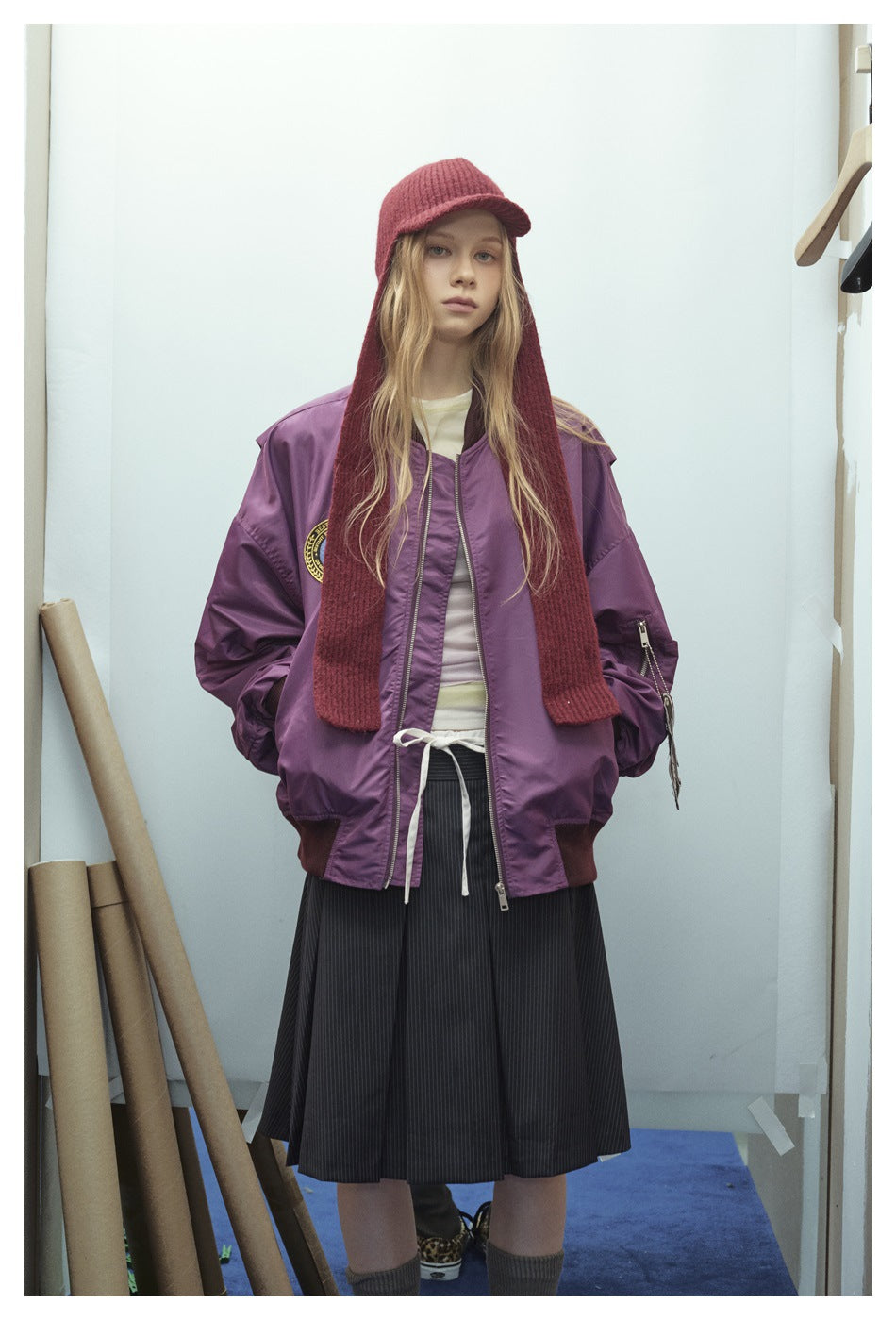 Model wearing a purple zip-front bomber jacket with a chest patch, burgundy knit hood scarf, and black pleated skirt.