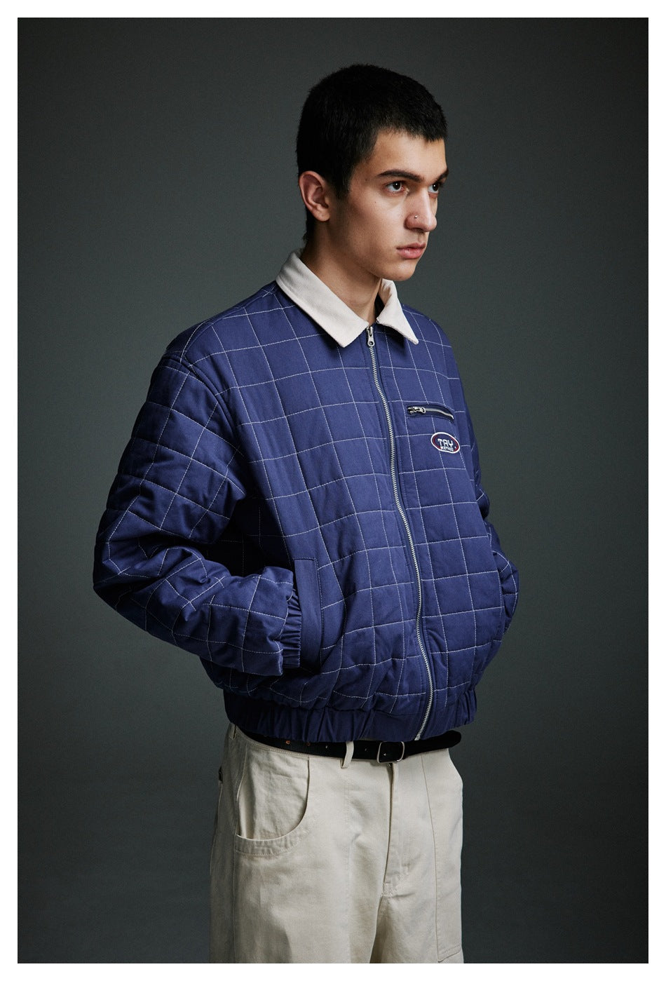 Model wearing a navy quilted jacket with white grid stitching, cream collar, zip front and chest pocket.