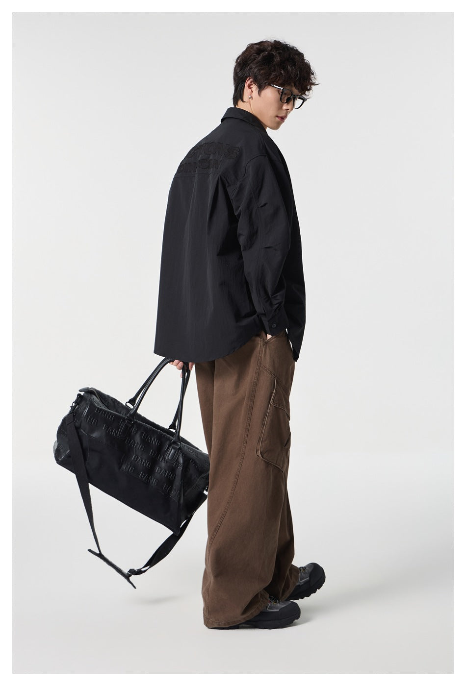 Model seen from the back wearing a black shirt with tonal embroidered back lettering, brown pants, holding a black bag