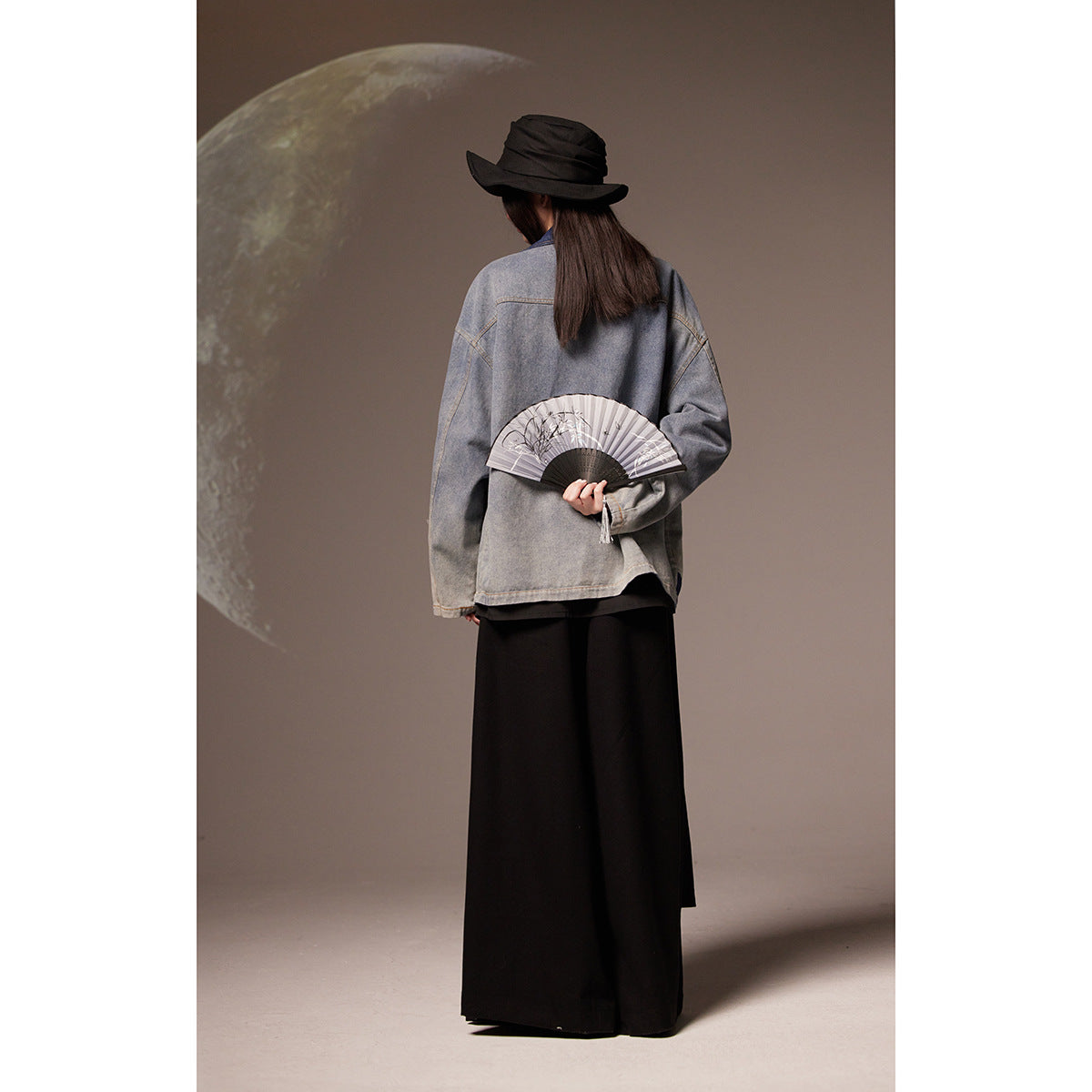 Back view of a model wearing a gray denim jacket with a long black skirt and black hat.