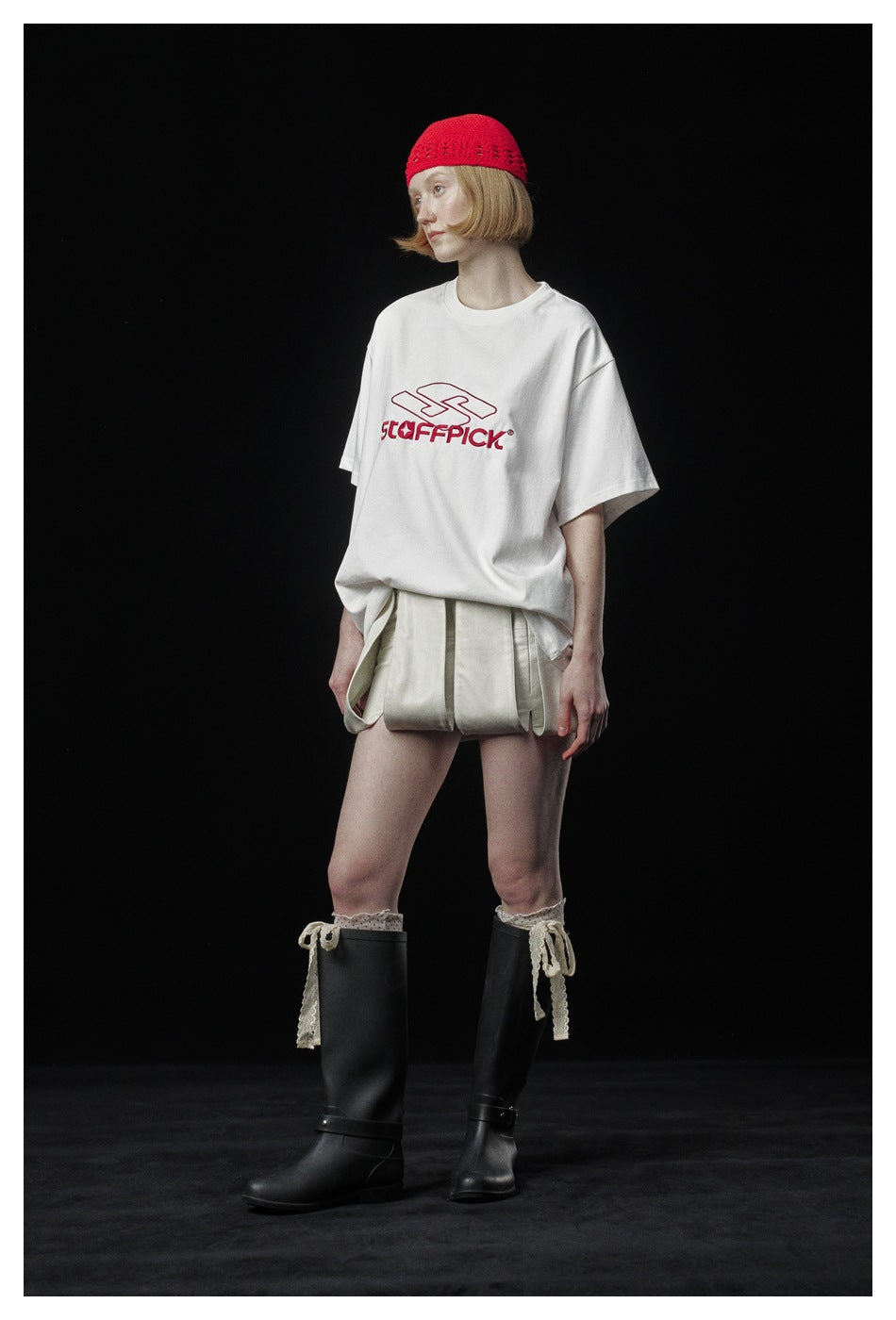 Three-quarter view of a model in a white graphic T-shirt, pale pleated shorts, a red knit cap, and black boots.