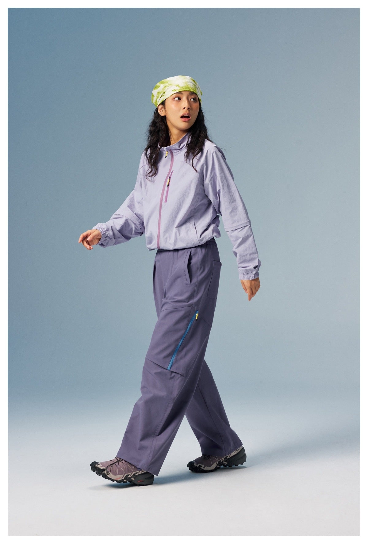 Model walking in a pale lavender zip jacket and slate-purple cargo pants with a blue-trimmed thigh pocket.