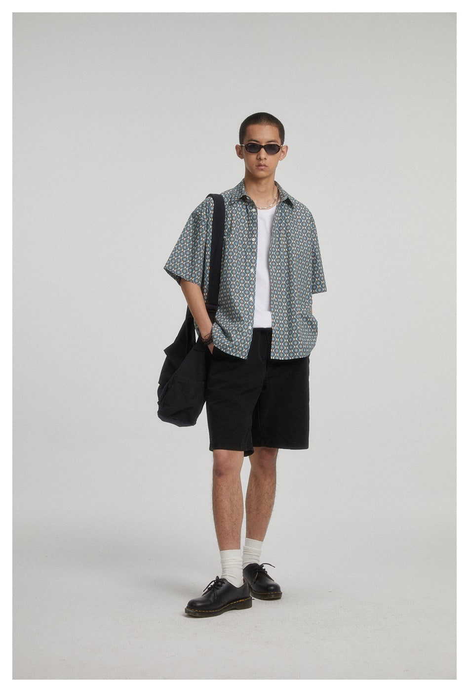Model wearing an open blue patterned short-sleeve shirt over a white top with black shorts.