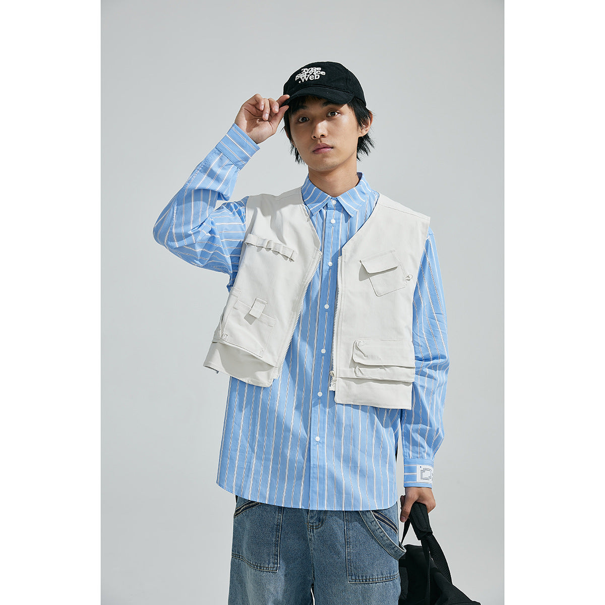 Model wearing a pale utility vest over a blue striped shirt, with jeans and a black cap.