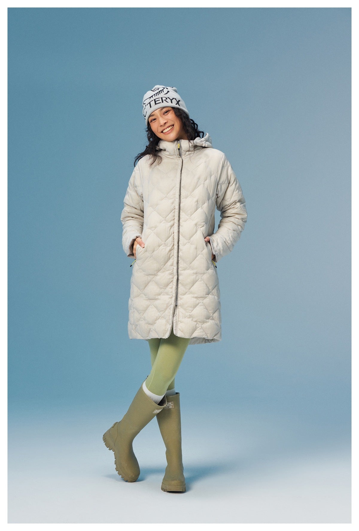 Model wearing a light cream diamond-quilted hooded coat zipped closed, with green leggings and olive knee-high boots.