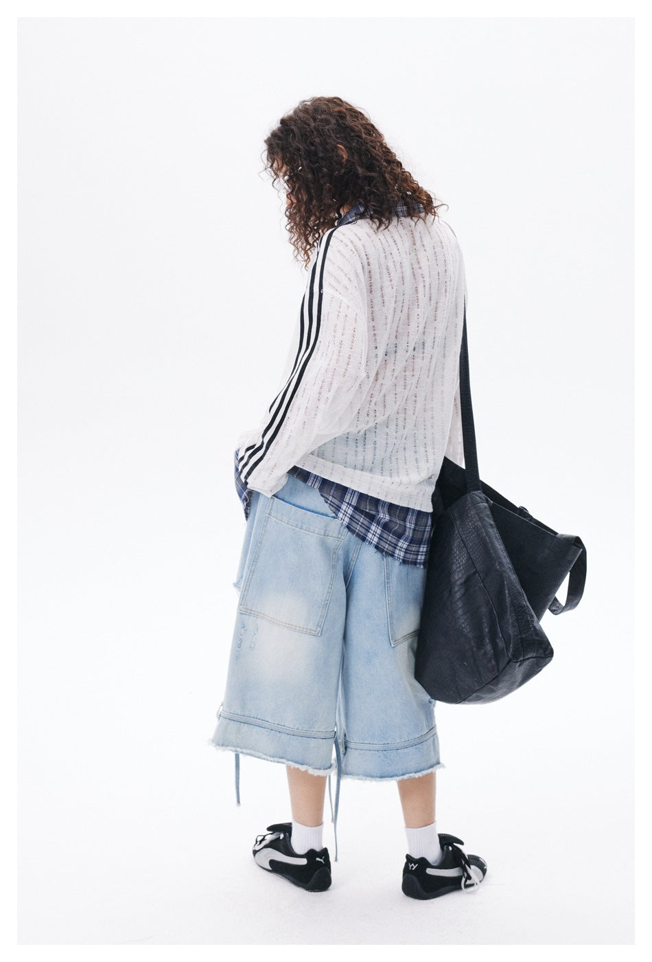 Rear view of a model wearing a white open-knit striped top and light denim shorts with a black shoulder bag.