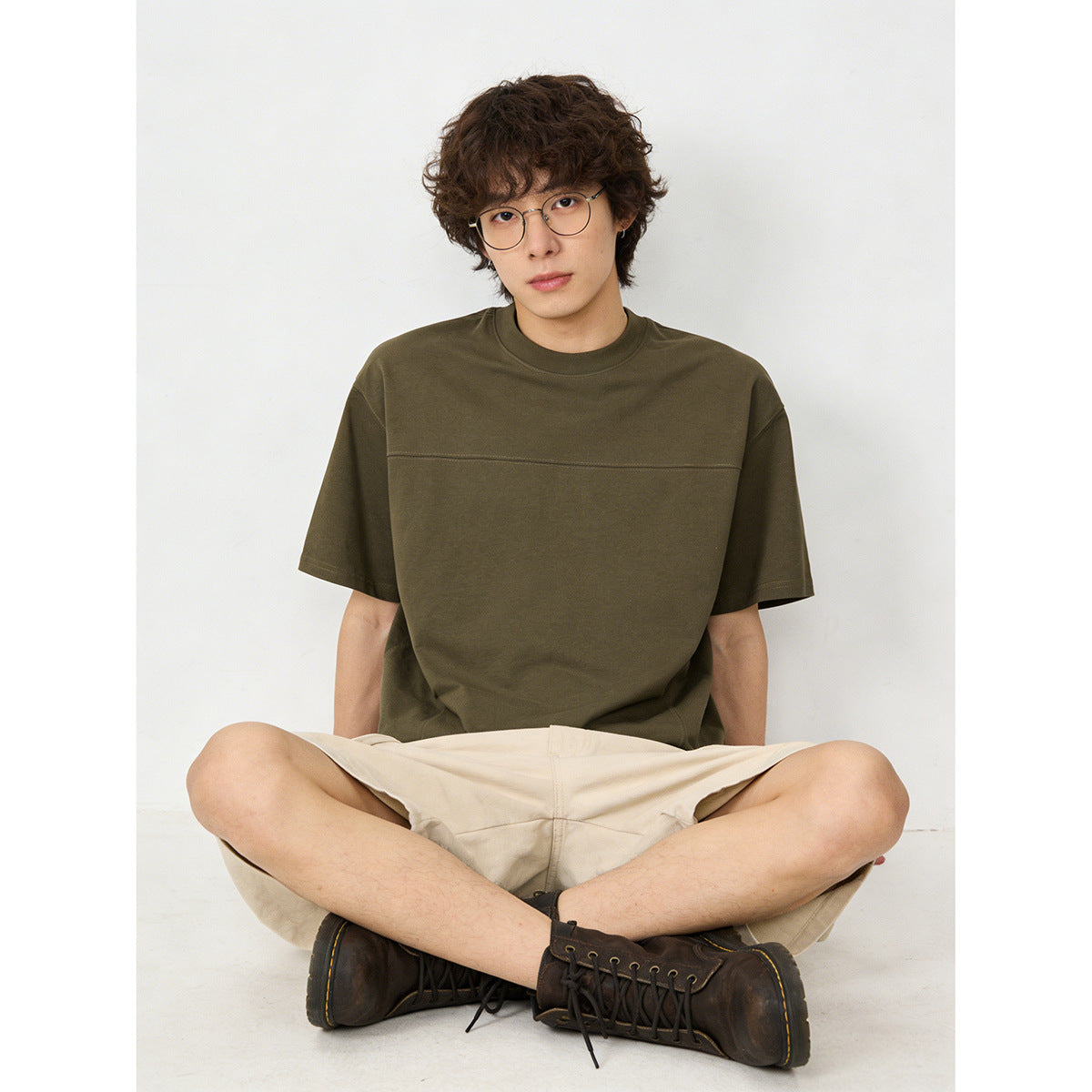 Seated model wearing an olive-brown oversized short-sleeve T-shirt with a horizontal chest seam.