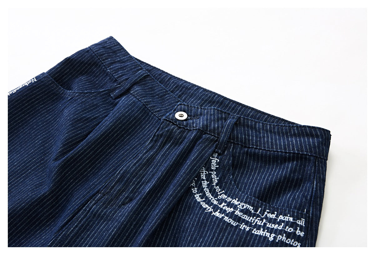 Angled close-up of the navy pinstriped shorts’ front waistband, button, pocket, and white script embroidery.