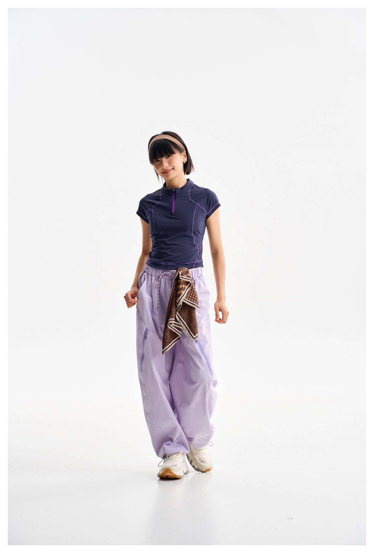 Model wearing a navy short-sleeve zip-neck top with purple piping and loose lavender pants, with a patterned scarf at the