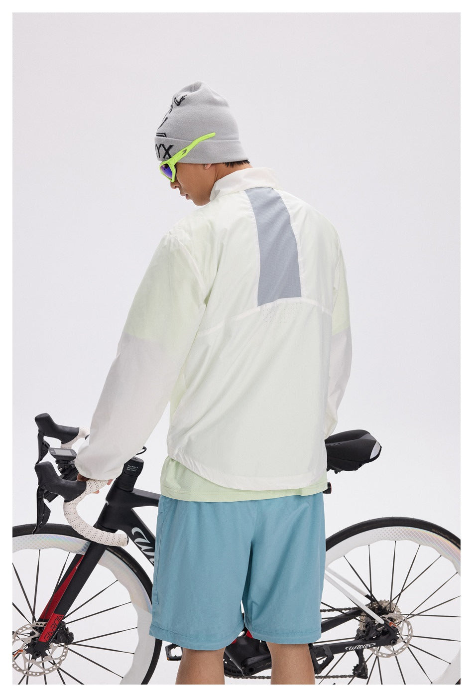 Rear view of a cyclist wearing a white windbreaker with a gray back panel and blue shorts.