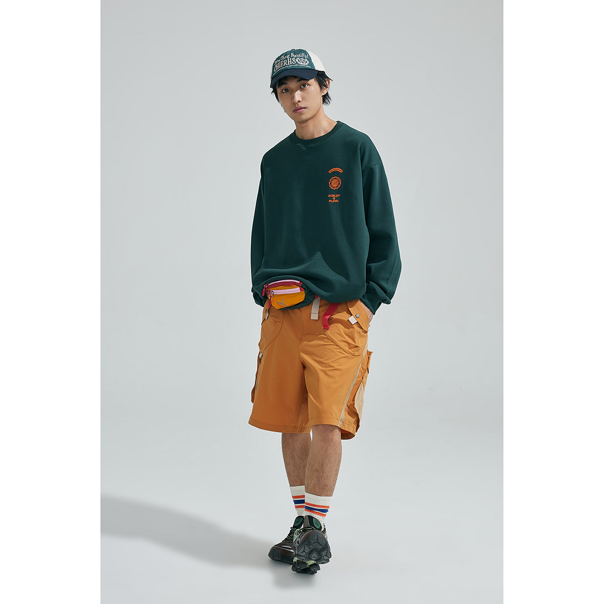 Front view of a model in a dark green sweatshirt with orange chest print and ochre cargo shorts.