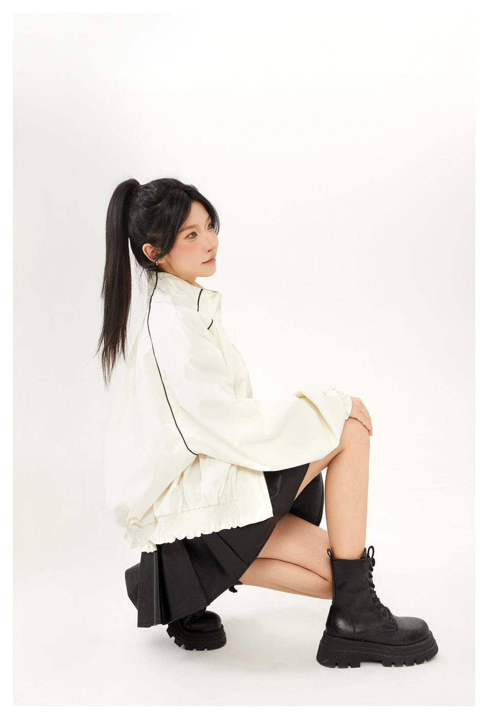 Model seated in an ivory high-collar jacket with black piping and gathered cuffs, paired with a black skirt and boots.