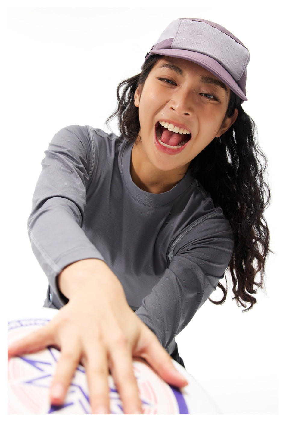 Close-up of a model wearing a gray long-sleeve top and lavender cap, reaching toward a flying disc.