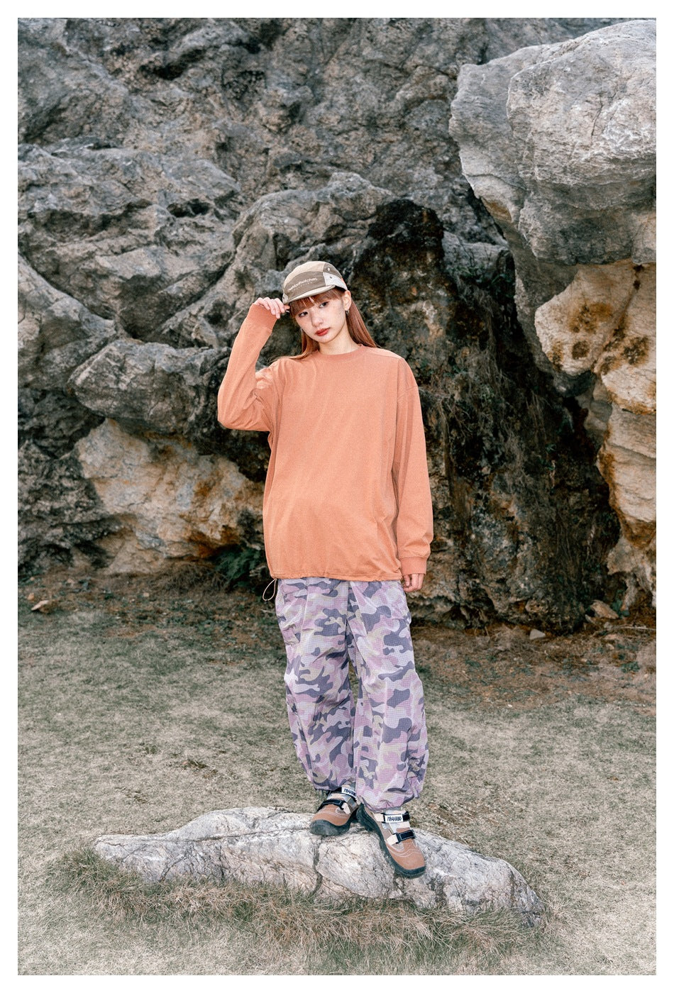 Woman outdoors in a peach long-sleeve shirt and loose lavender camouflage trousers, standing among rocks.